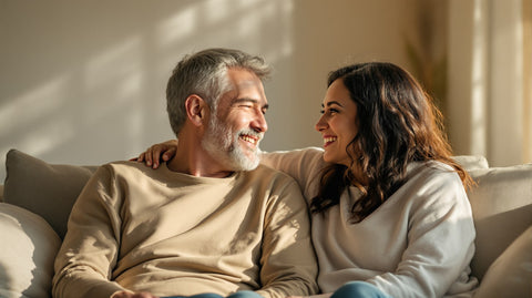 A man and his wife sitting close together on a sofa at home in warm morning light, smiling at each other
